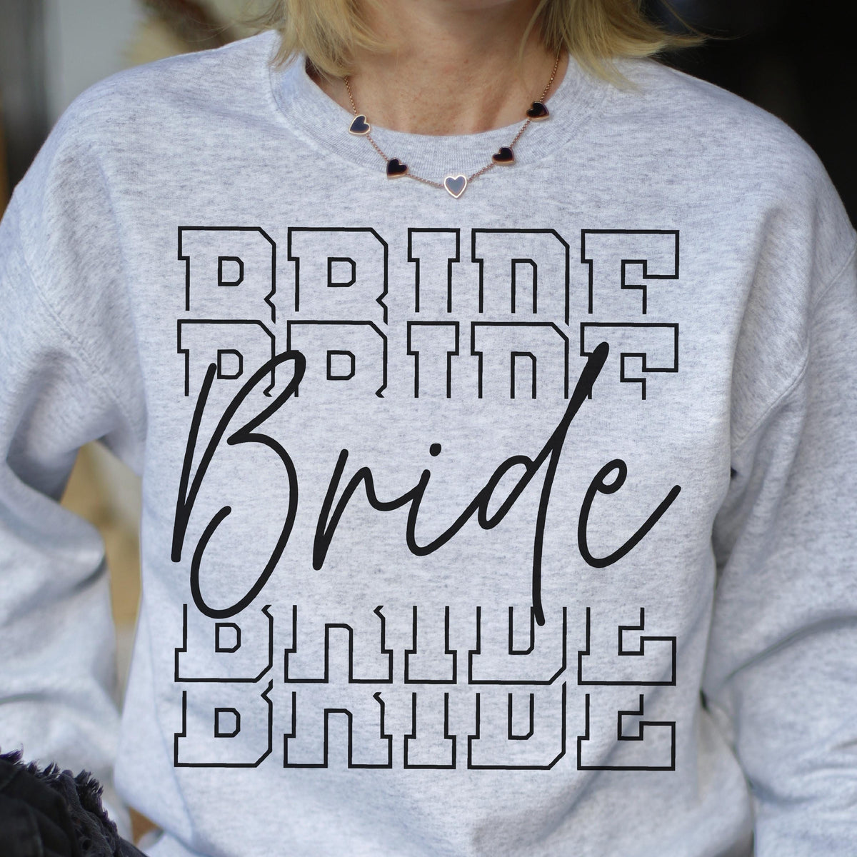 May include: A light gray crewneck sweatshirt featuring the word BRIDE printed in a black block font four times, with the word Bride written in a large black cursive script layered over the center of the block text.