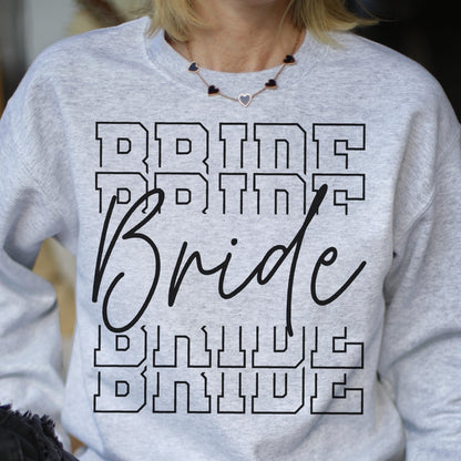 May include: A light gray crewneck sweatshirt featuring the word BRIDE printed in a black block font four times, with the word Bride written in a large black cursive script layered over the center of the block text.