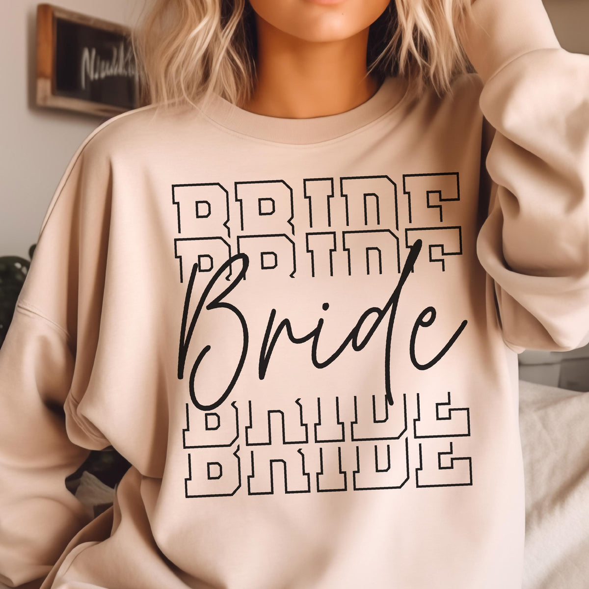 May include: A beige crewneck sweatshirt featuring the word Bride printed in a black cursive script font across the center, layered over and under the word BRIDE repeated four times in a black outlined block letter varsity style font.