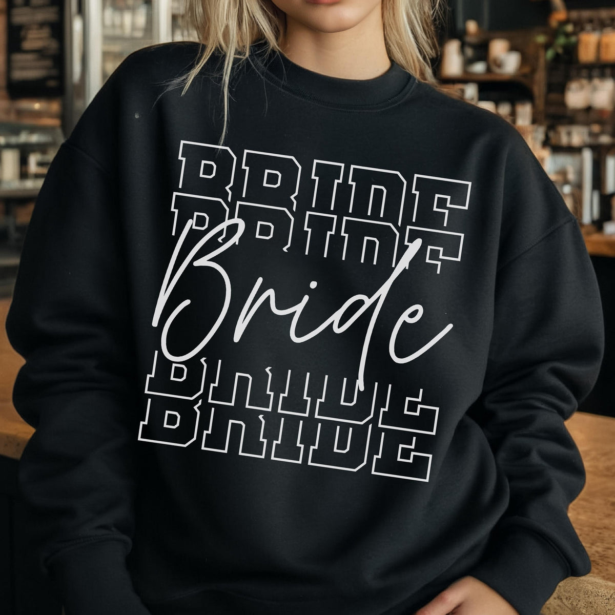 May include: A black long-sleeve crewneck sweatshirt featuring the word BRIDE printed in a white block varsity font above and below the word Bride written in a white cursive script, creating a layered text design across the chest.