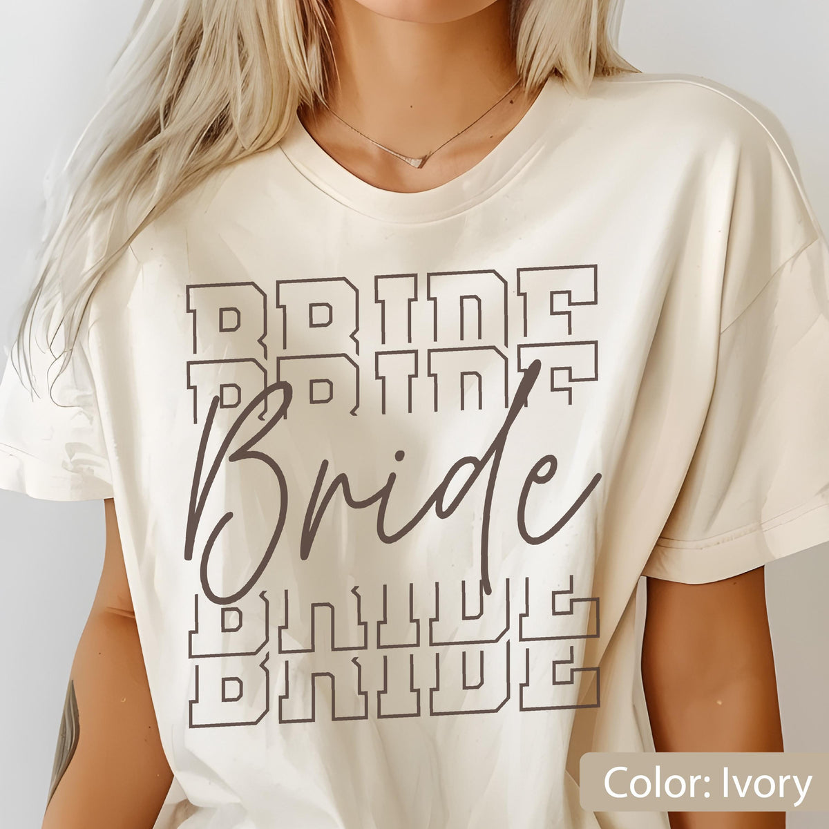 May include: Ivory-colored t-shirt with the word "Bride" in a stacked, outlined font. The word "Bride" is also written in a cursive font over the stacked text. The shirt is a relaxed fit.