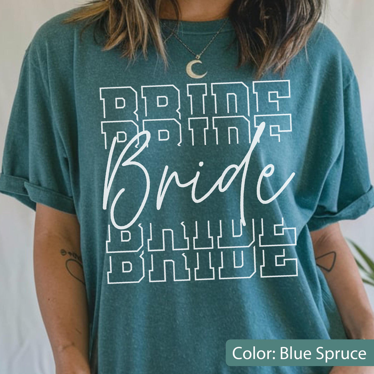 May include: Blue spruce t-shirt with the word "Bride" in a white, layered design. The word "Bride" is written in a cursive font, with the word "Bride" repeated in a block letter font above and below.