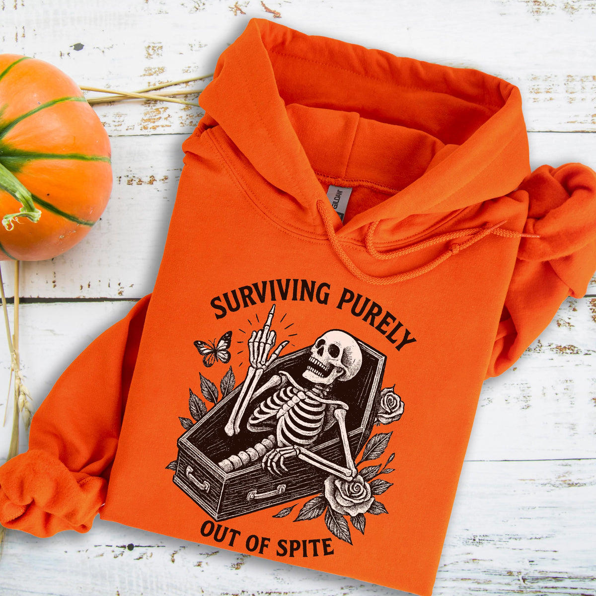 May include: Orange hoodie featuring a skeleton in a coffin with the text "SURVIVING PURELY OUT OF SPITE." The design includes a butterfly and roses. The hoodie is made of a soft material.