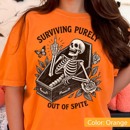 May include: An orange t-shirt featuring a black graphic print of a skeleton lying in a coffin, raising its middle finger. The text "SURVIVING PURELY" is arched above, and "OUT OF SPITE" is below. The design includes a butterfly and roses.