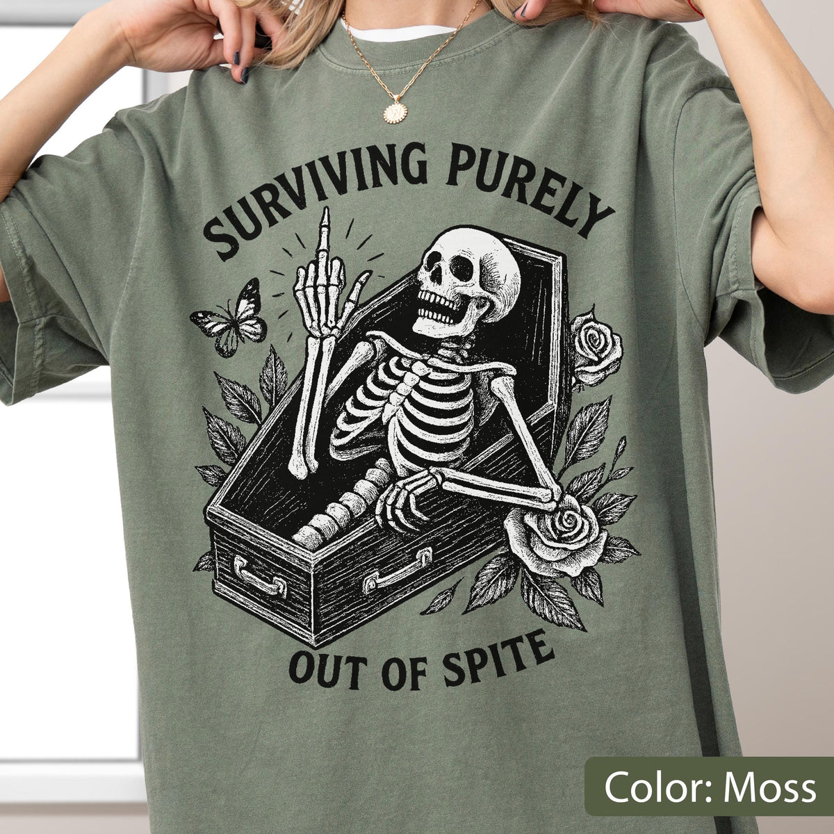 May include: A moss green t-shirt featuring a black graphic print of a skeleton lying in a coffin. The skeleton is raising its middle finger. The text "SURVIVING PURELY" is arched above the coffin, and "OUT OF SPITE" is printed below, with roses and a butterfly.