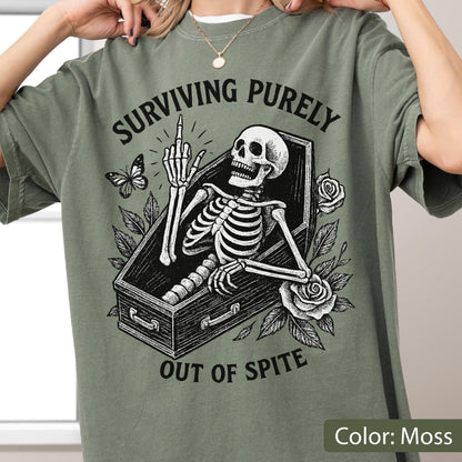 May include: A moss green t-shirt featuring a black graphic print of a skeleton lying in a coffin. The skeleton is raising its middle finger. The text "SURVIVING PURELY" is arched above the coffin, and "OUT OF SPITE" is printed below, with roses and a butterfly.