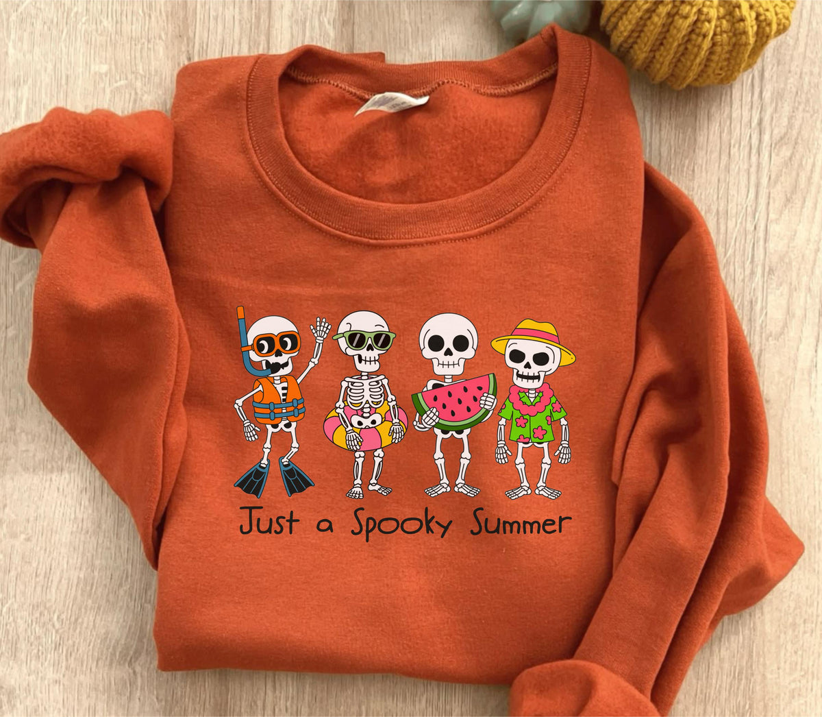 May include: An orange sweatshirt featuring a graphic of four skeleton characters in summer attire. One skeleton wears a snorkel, another sunglasses and a floatie, a third holds watermelon, and the last wears a straw hat. The text "Just a Spooky Summer" is below.