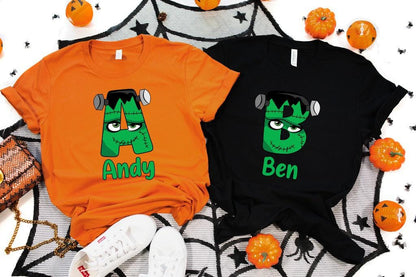 May include: Two Halloween-themed t-shirts, one orange and one black, each featuring a Frankenstein's monster letter design. The orange shirt has the letter "A" with the name "Andy" below. The black shirt has the letter "B" with the name "Ben".