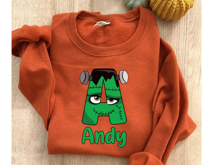 May include: An orange sweatshirt with a green Frankenstein's monster-themed letter "A" graphic. The graphic includes the name "Andy" in green. The sweatshirt is folded on a wooden surface.