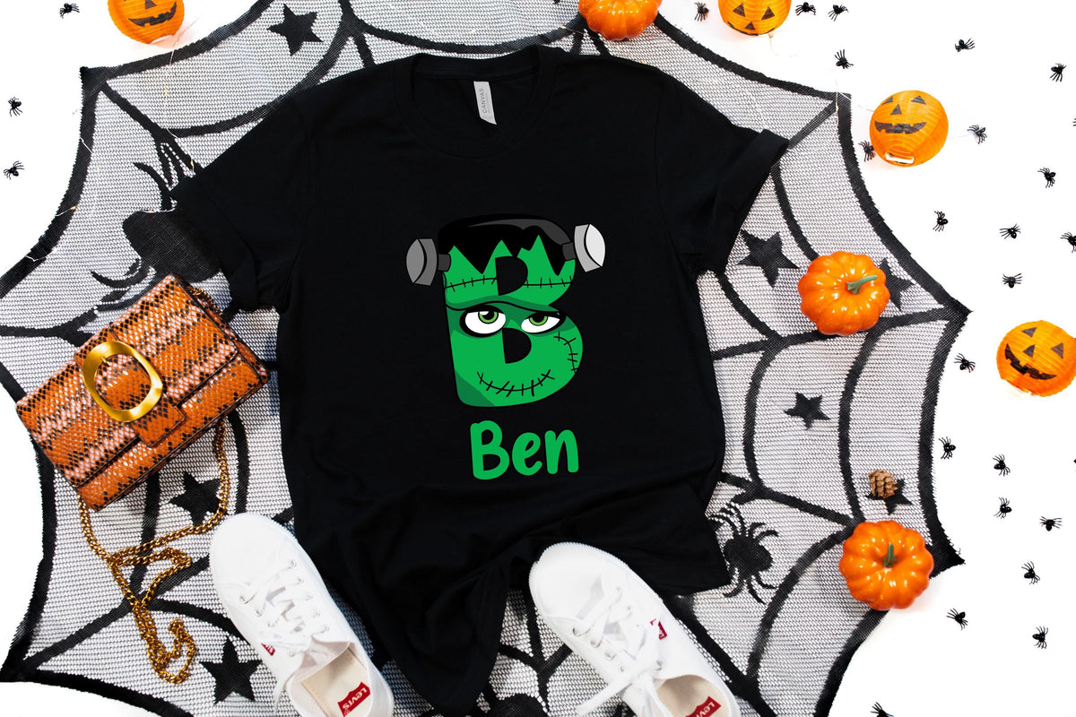 May include: Black t-shirt with a green Frankenstein monster-themed letter "B" and the name "Ben" in green. The shirt is surrounded by Halloween decorations, including pumpkins and a spiderweb.