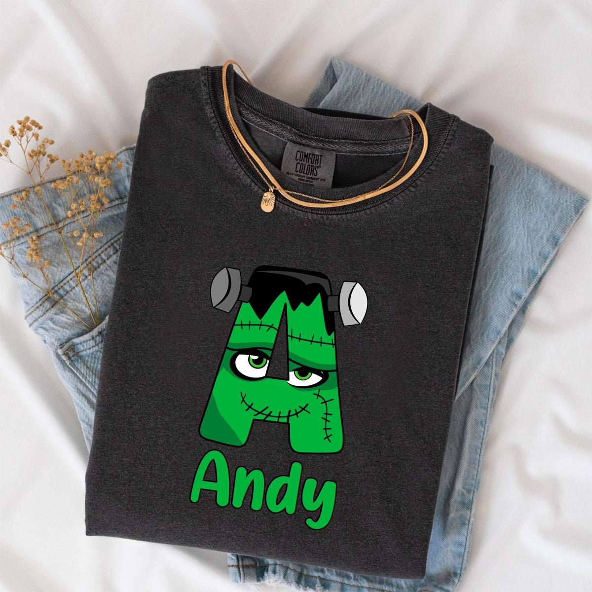 May include: A dark gray t-shirt with a green Frankenstein monster letter "A" design and the name "Andy" in green. The shirt is folded on top of a pair of blue jeans and a gold necklace.
