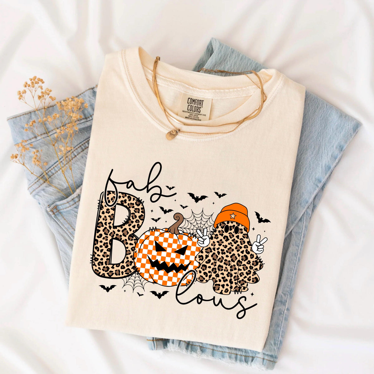 May include: A cream-colored t-shirt featuring a graphic with the text "fab" and "lous" in black script. The design includes a leopard print letter B, a checkered orange and white pumpkin, and a leopard print ghost wearing an orange beanie.