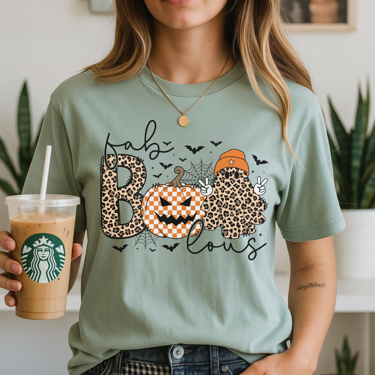 May include: A sage green t-shirt featuring a graphic print with the text "fab-lous" in black script. The design includes a leopard print letter B, an orange and white checkered pumpkin, and a leopard print ghost wearing an orange beanie.