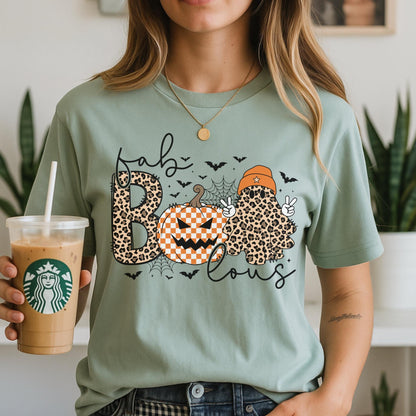 May include: A sage green t-shirt featuring a graphic print with the text "fab-lous" in black script. The design includes a leopard print letter B, an orange and white checkered pumpkin, and a leopard print ghost wearing an orange beanie.