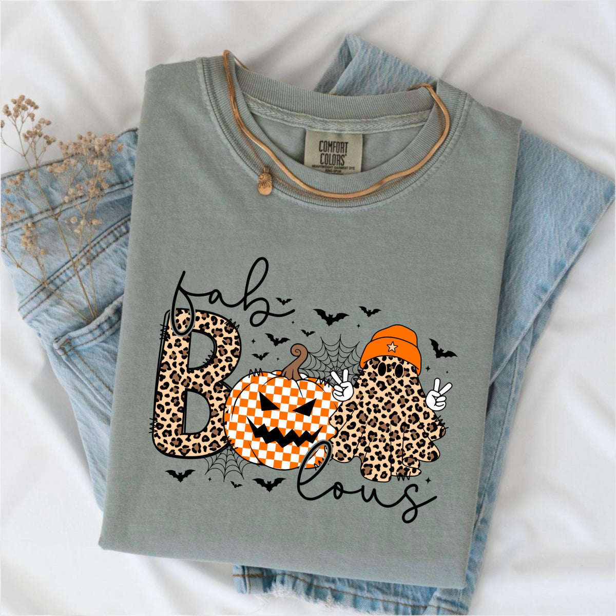 May include: A sage green t-shirt with a Comfort Colors label at the neck. The graphic features the text "fab-BOO-lous" with a leopard print letter B, a checkered orange and white pumpkin, and a leopard print ghost wearing an orange beanie.