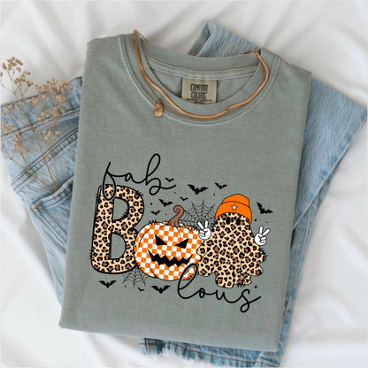 May include: A sage green t-shirt with a Comfort Colors label at the neck. The graphic features the text "fab-BOO-lous" with a leopard print letter B, a checkered orange and white pumpkin, and a leopard print ghost wearing an orange beanie.