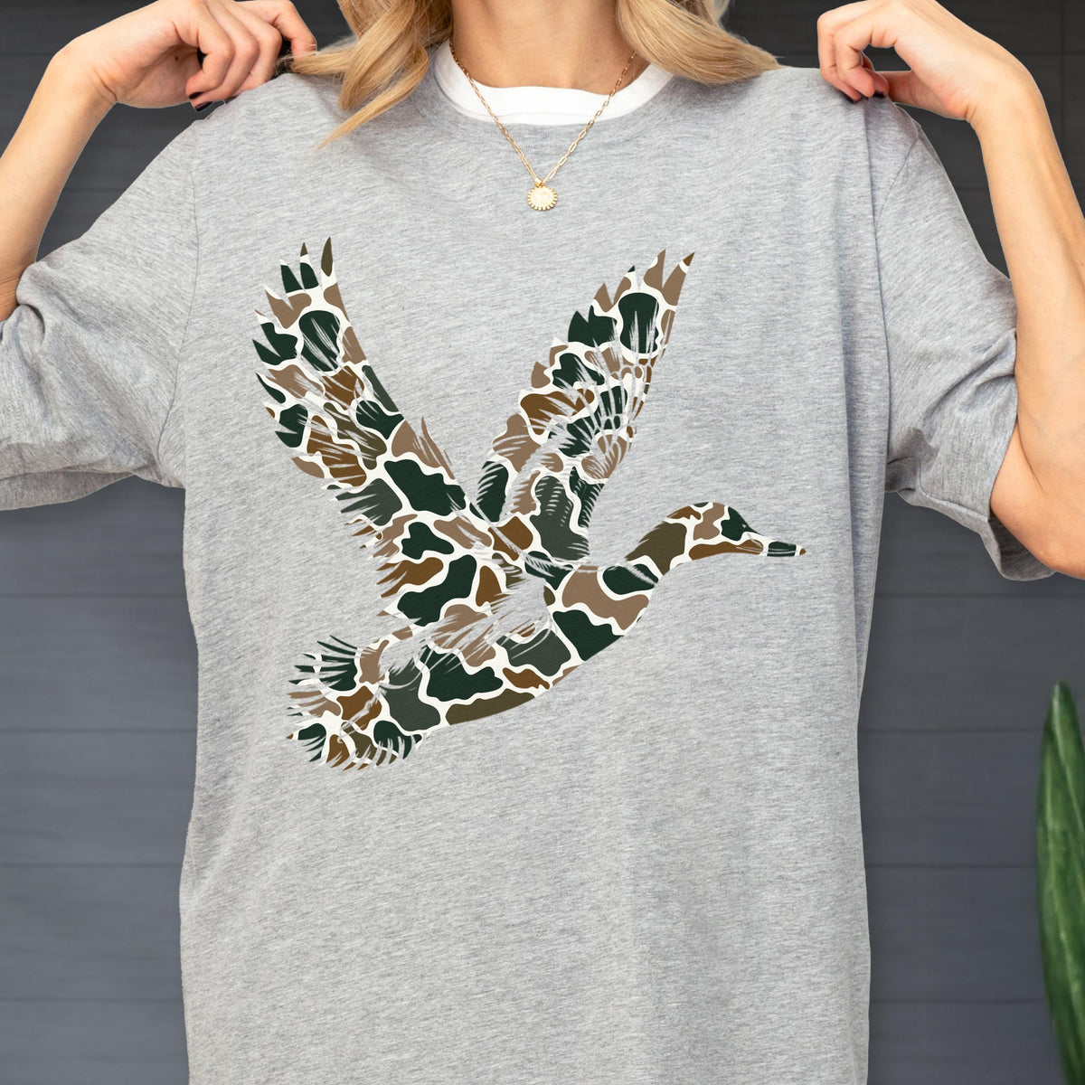 May include: A heather gray short-sleeve t-shirt featuring a large graphic of a flying duck. The duck is filled with a camouflage pattern consisting of dark green, brown, and tan organic shapes. The shirt has a crew neckline and a relaxed fit.