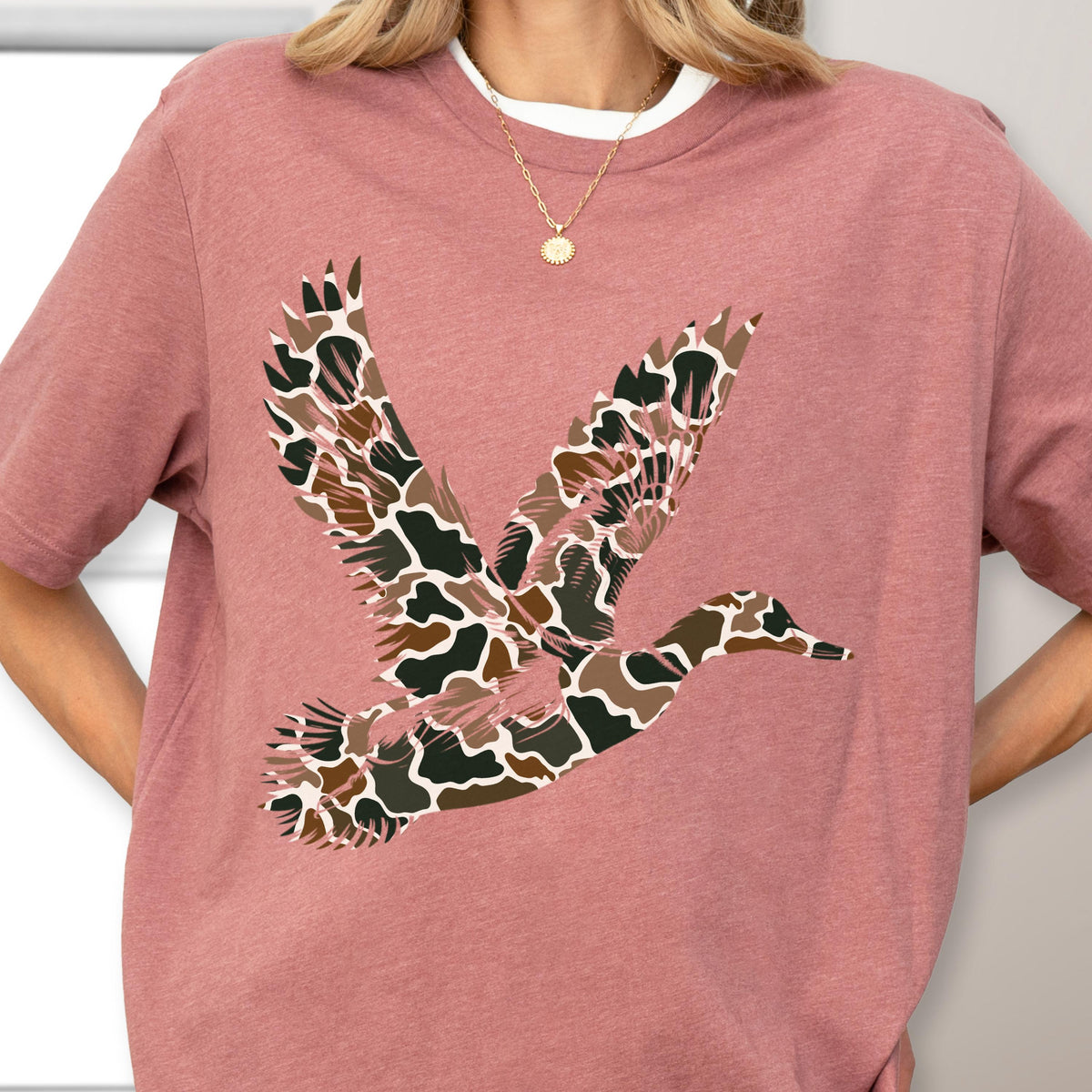 May include: A dusty rose short-sleeve t-shirt featuring a graphic of a flying duck. The duck is filled with a brown, black, and white camouflage pattern. The shirt has a crew neckline and is worn with a thin gold chain necklace with a small pendant.