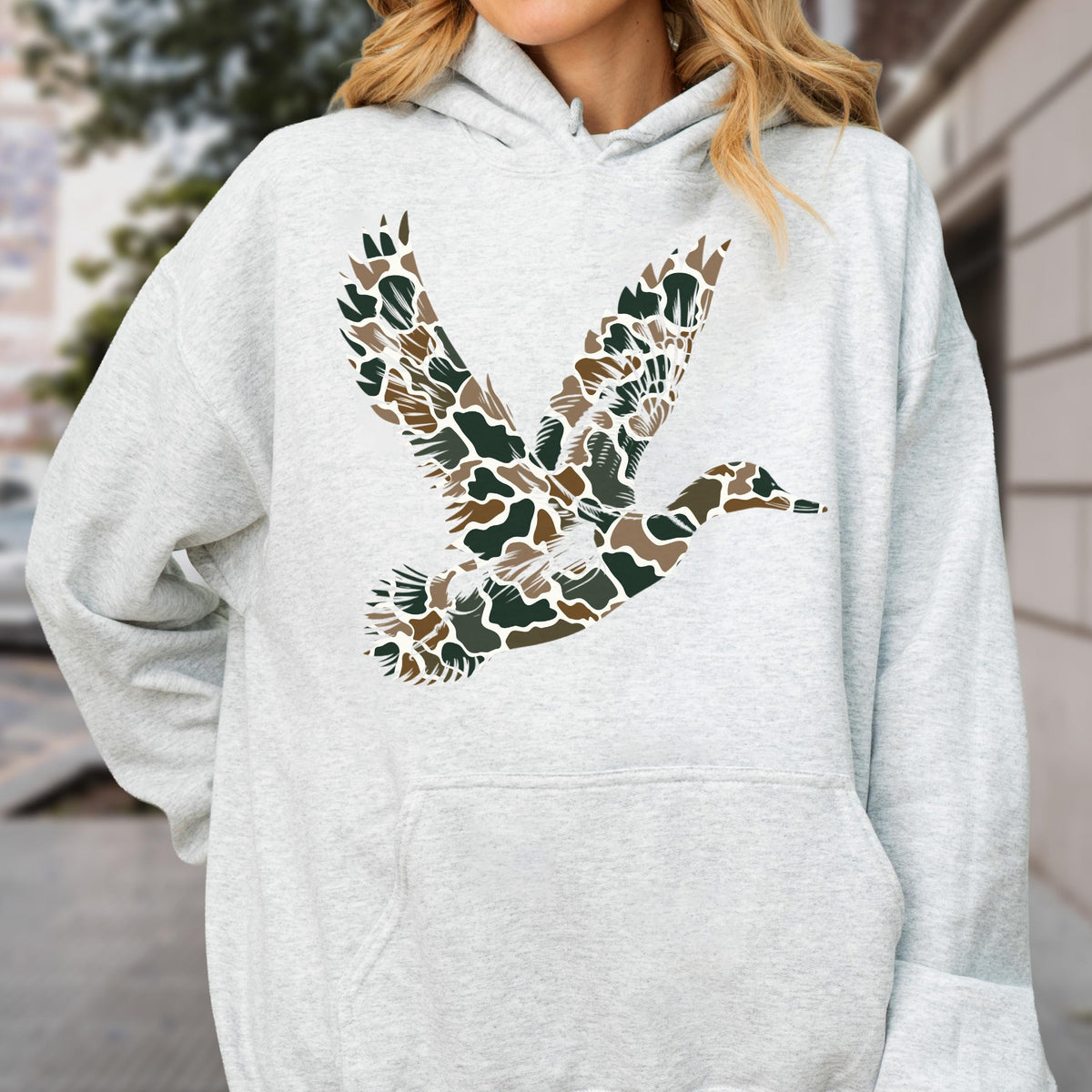 May include: A light gray heathered hooded sweatshirt featuring a large graphic of a flying duck on the chest. The duck silhouette is filled with a camouflage pattern consisting of brown, dark green, and tan organic shapes on a white background.