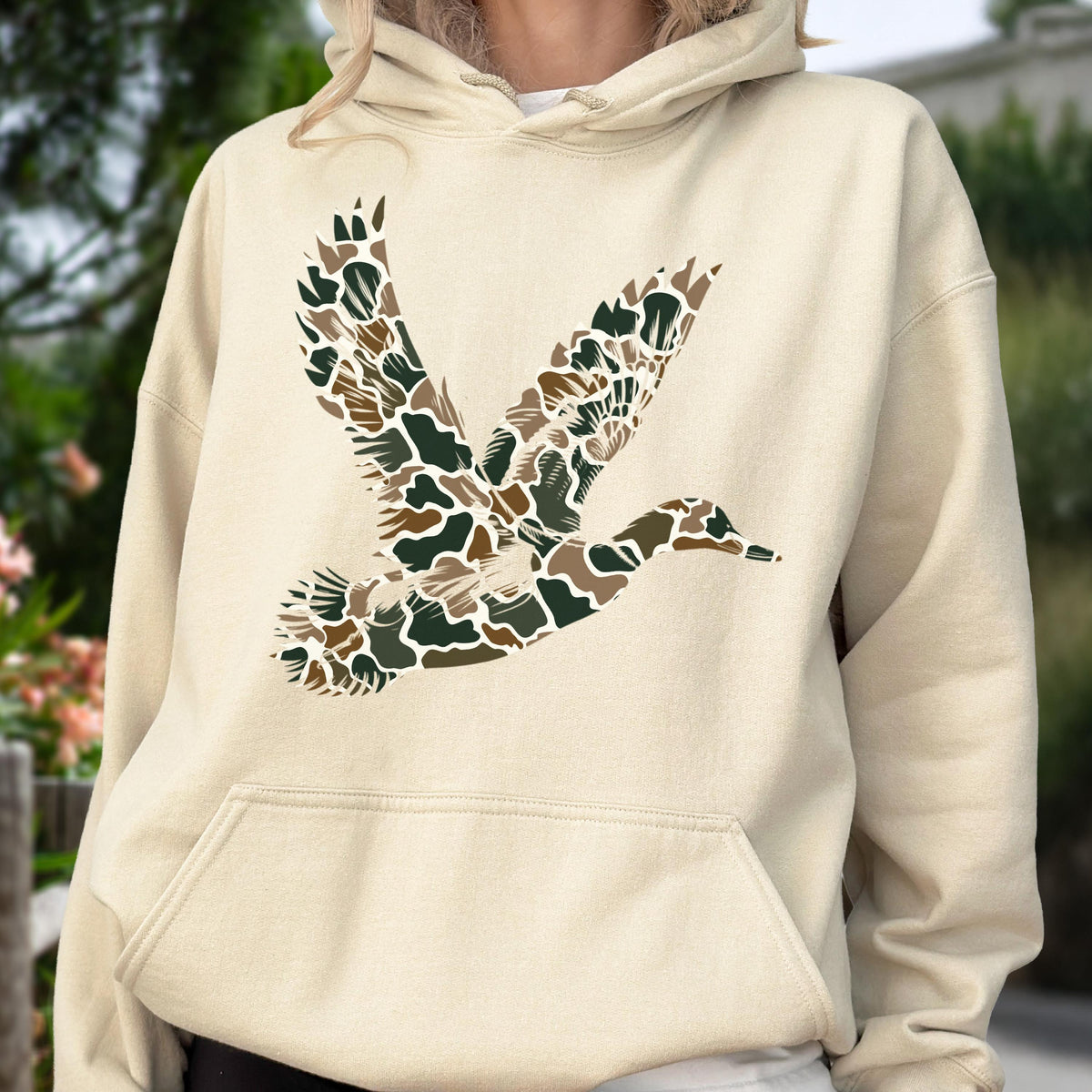 May include: A cream-colored hooded sweatshirt featuring a large graphic of a flying duck on the chest. The duck silhouette is filled with a camouflage pattern consisting of various shades of brown, dark green, and tan shapes.