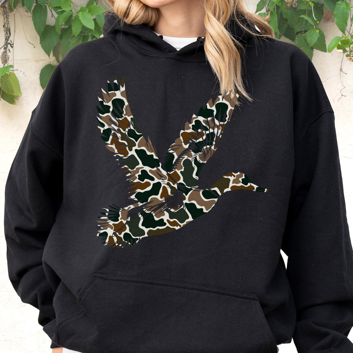 May include: A black hooded sweatshirt featuring a large graphic of a flying duck on the chest. The duck silhouette is filled with a camouflage pattern consisting of brown, dark green, and black organic shapes. The hoodie includes a front pocket.