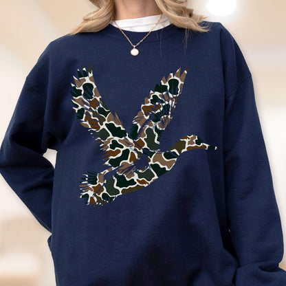 May include: A navy blue crewneck sweatshirt featuring a large graphic of a flying duck filled with a brown, tan, and dark green camouflage pattern. The sweatshirt has long sleeves and a ribbed neckline, worn with a simple gold chain necklace.