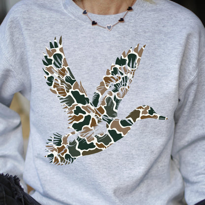 May include: A light gray crewneck sweatshirt featuring a large graphic of a flying duck. The duck is filled with a camouflage pattern consisting of brown, dark green, and white organic shapes. The sweatshirt has long sleeves and a ribbed neckline.