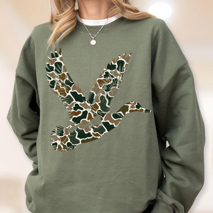 May include: An olive green crewneck sweatshirt featuring a large graphic of a flying duck. The duck is filled with a camouflage pattern consisting of brown, tan, and dark green shapes. The sweatshirt has long sleeves and a ribbed collar.
