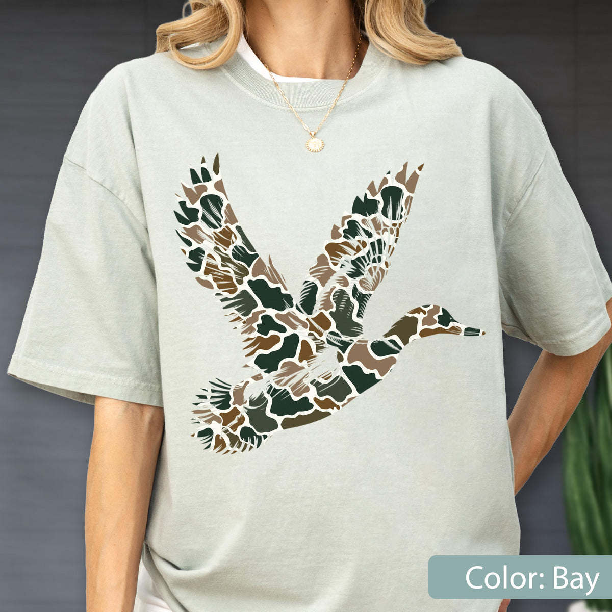 May include: A light sage green short-sleeve t-shirt featuring a large graphic of a flying duck. The duck silhouette is filled with a camouflage pattern consisting of dark green, brown, and tan organic shapes. The shirt has a crew neckline.