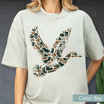 May include: A light sage green short-sleeve t-shirt featuring a large graphic of a flying duck. The duck silhouette is filled with a camouflage pattern consisting of dark green, brown, and tan organic shapes. The shirt has a crew neckline.