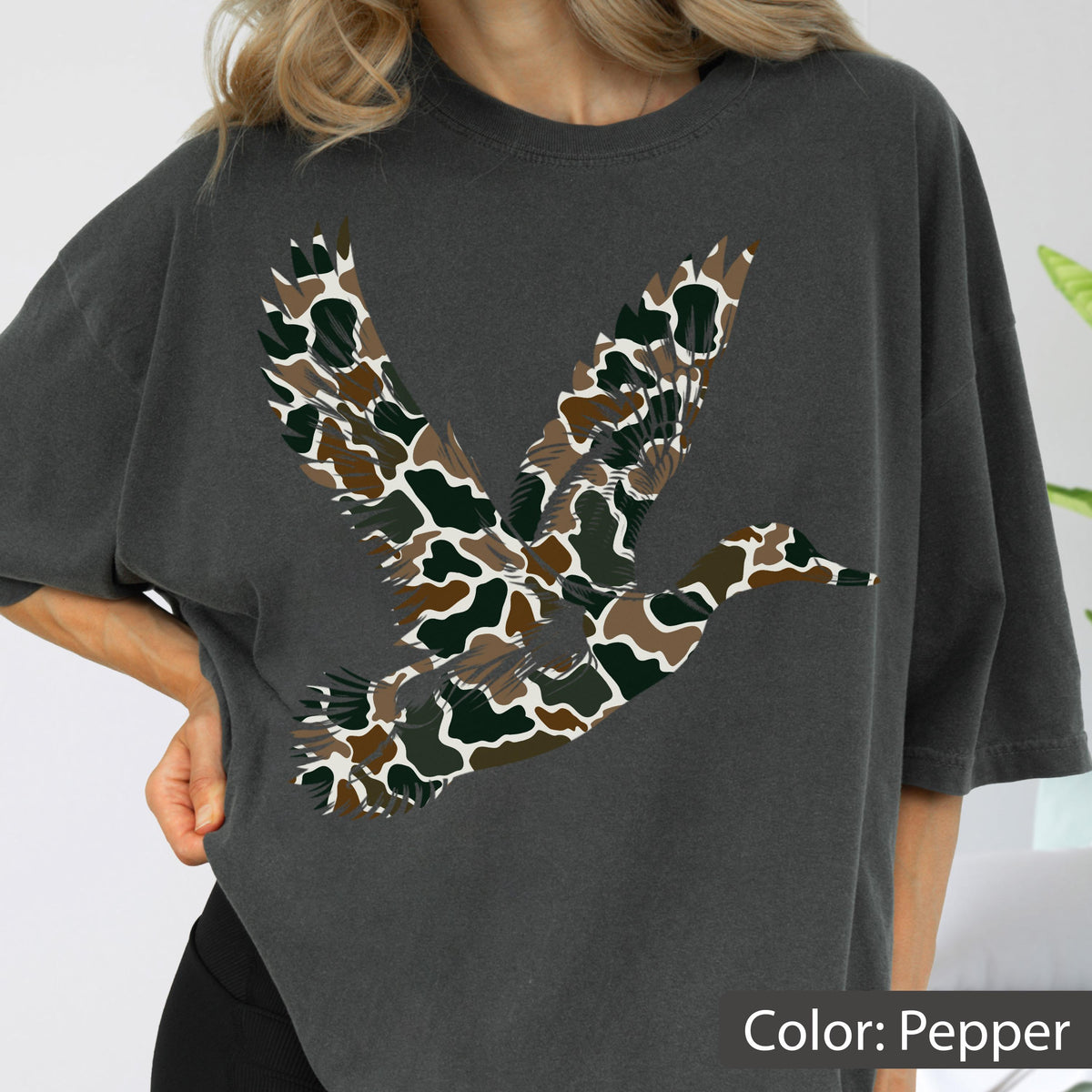 May include: A charcoal gray short-sleeve t-shirt featuring a large graphic of a flying duck. The duck silhouette is filled with a camouflage pattern consisting of brown, dark green, and black organic shapes. The shirt has a crew neckline.