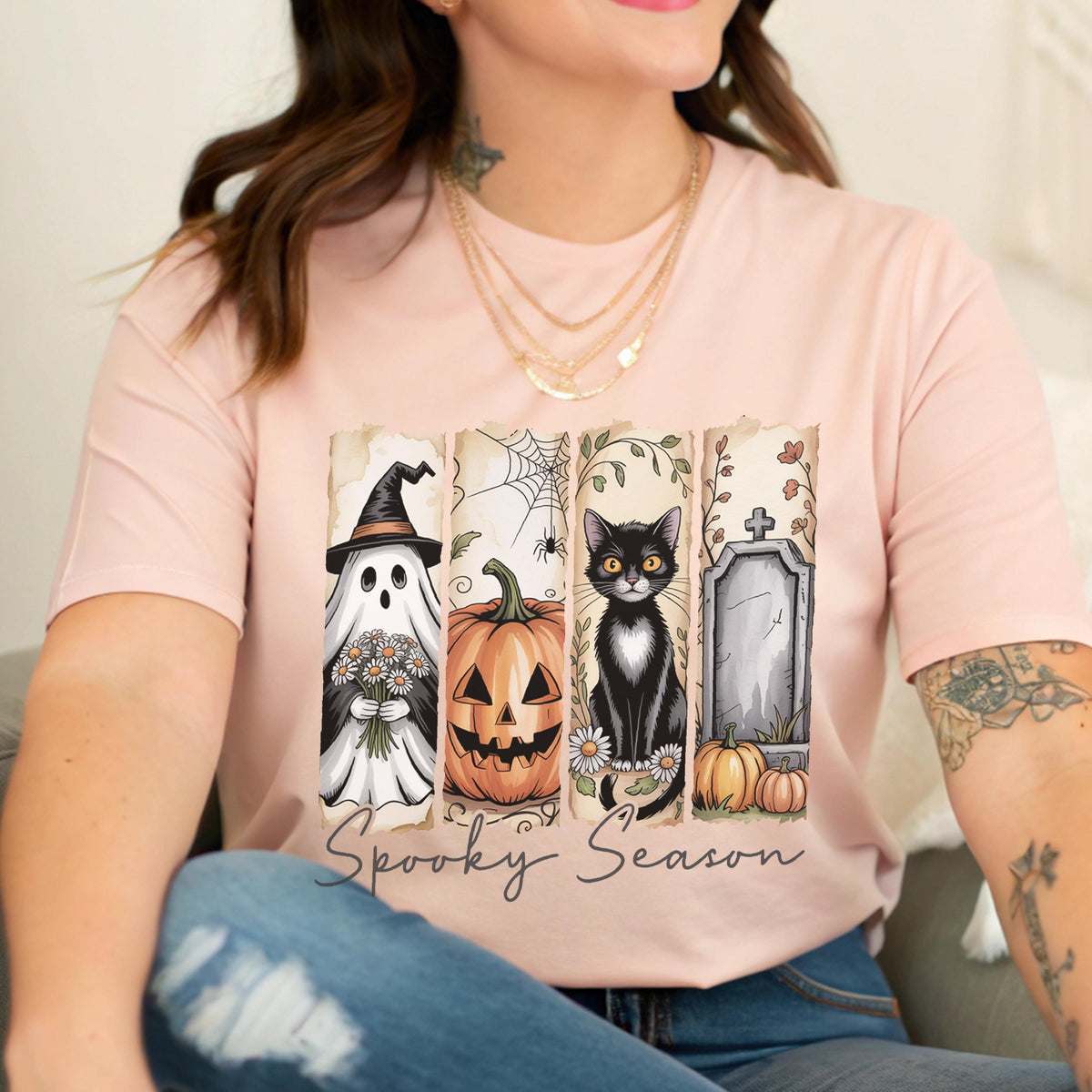 May include: A light pink t-shirt featuring a graphic print of four vertical panels. The panels display a ghost in a witch hat holding flowers, a jack-o'-lantern, a black cat with white markings, and a tombstone. The text Spooky Season is below.