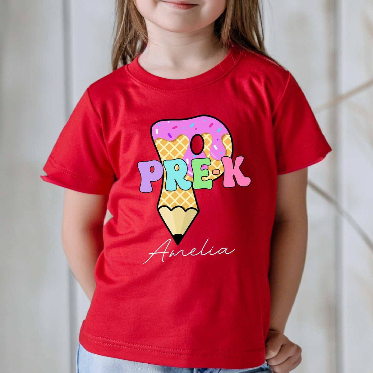 May include: A red t-shirt featuring a colorful graphic for Pre-K. The design includes a donut, a pencil, and the name "Amelia" in script. The shirt is made of a soft material.