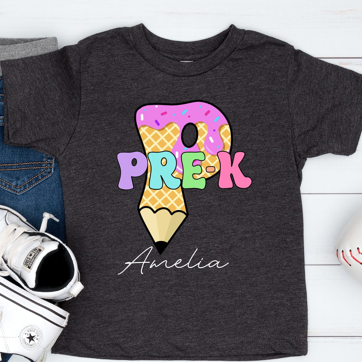 May include: Dark gray t-shirt featuring a colorful graphic for Pre-K. The design includes the text "PRE-K" in pastel colors, a donut, and a pencil. The name "Amelia" is written in cursive below the graphic.