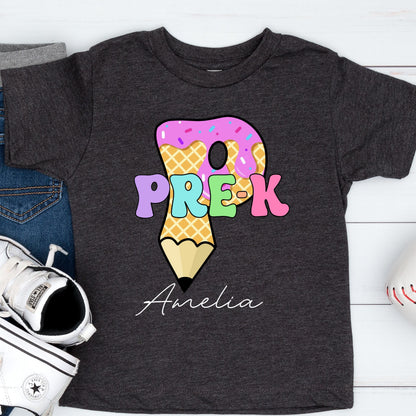 May include: Dark gray t-shirt featuring a colorful graphic for Pre-K. The design includes the text "PRE-K" in pastel colors, a donut, and a pencil. The name "Amelia" is written in cursive below the graphic.