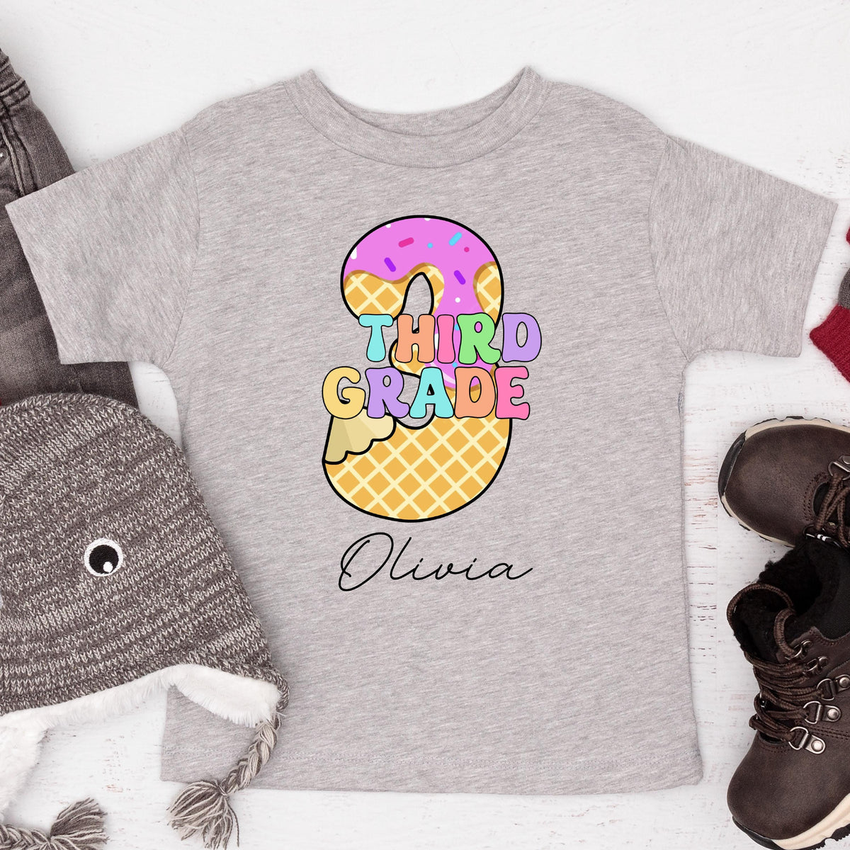 May include: A light gray t-shirt featuring a colorful graphic of a waffle cone and donut shaped like the number 3. The words "THIRD GRADE" are in pastel colors, with the name "Olivia" below in cursive.