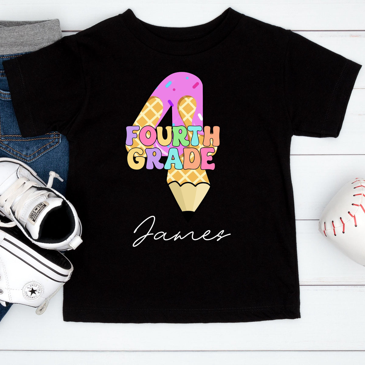 May include: Black t-shirt with a colorful "4" design, featuring a waffle pattern and the words "Fourth Grade." A pencil shape forms the bottom of the "4." The name "James" is written in cursive below the design.