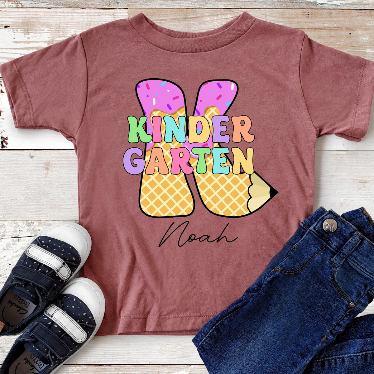 May include: A maroon short-sleeved t-shirt with the words "KINDERGARTEN" in colorful letters. The design includes a pencil and ice cream cone shapes. The name "Noah" is written in cursive below the design. Dark blue jeans and navy blue shoes are also visible.