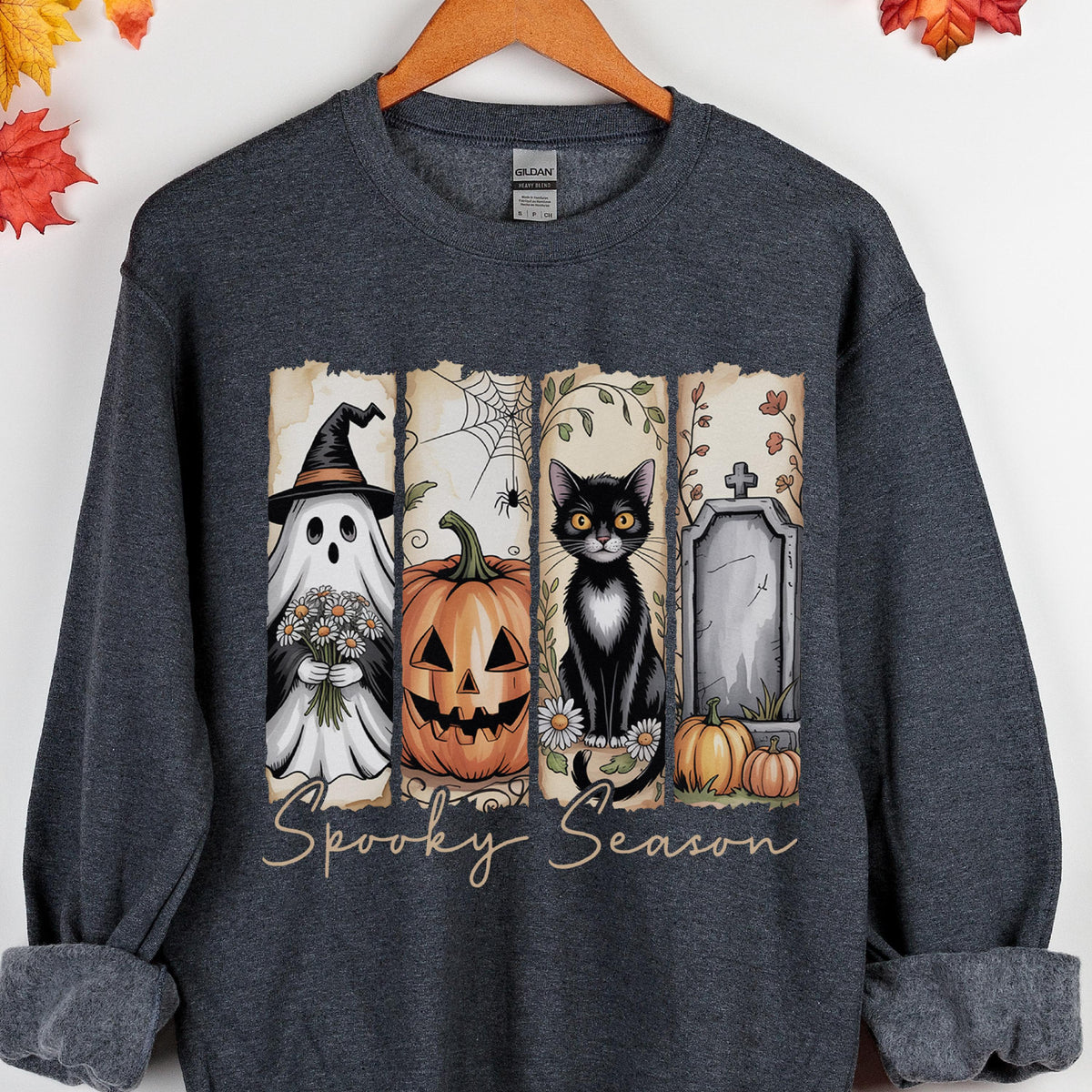 May include: A dark charcoal gray crewneck sweatshirt featuring a graphic print of four vertical panels. The panels display a ghost in a witch hat, a jack-o'-lantern, a black cat with daisies, and a tombstone. The text Spooky Season is printed below.