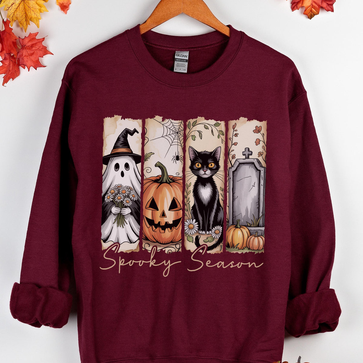 May include: A maroon long-sleeve sweatshirt featuring a graphic print of four vertical panels with a ghost holding flowers, a jack-o'-lantern, a black cat, and a tombstone. The text "Spooky Season" is printed in a cursive font below the panels.