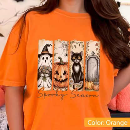 May include: Orange t-shirt featuring a Halloween-themed graphic. The design includes a ghost, jack-o'-lantern, black cat, and tombstone, with the words "Spooky Season" below. The shirt is a solid orange color.