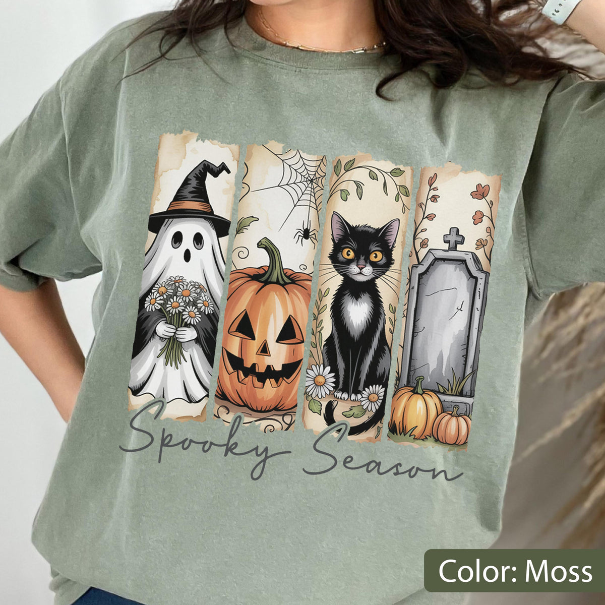 May include: Moss green t-shirt featuring a Halloween-themed graphic. The design includes a ghost, jack-o'-lantern, black cat, and tombstone. The words "Spooky Season" are written across the bottom of the graphic.