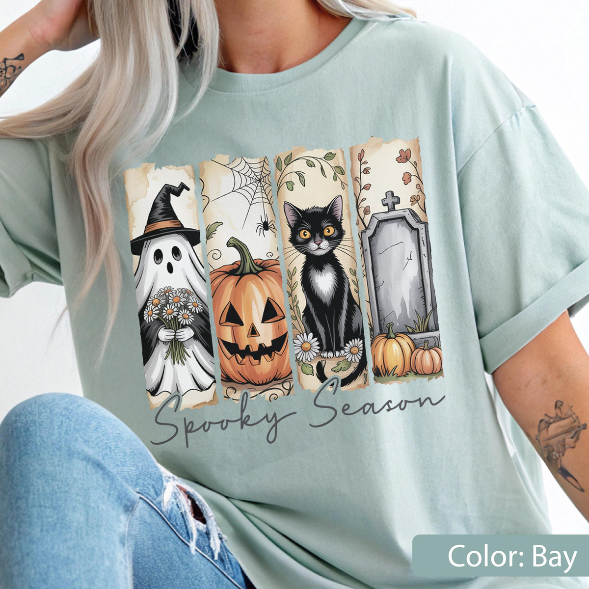 May include: A light green t-shirt featuring a Halloween-themed design. The design includes a ghost, jack-o'-lantern, black cat, and tombstone. The words "Spooky Season" are printed below the images. The shirt's color is "Bay".