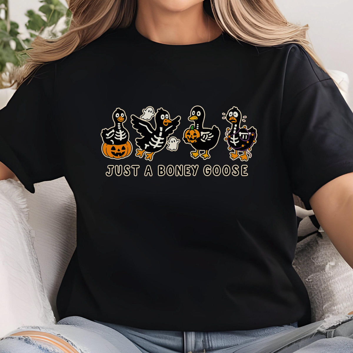 May include: Black t-shirt featuring a Halloween-themed graphic. The design includes four cartoon geese dressed in skeleton costumes, holding pumpkins and wearing ghost costumes. The text "JUST A BONEY GOOSE" is printed below the geese.