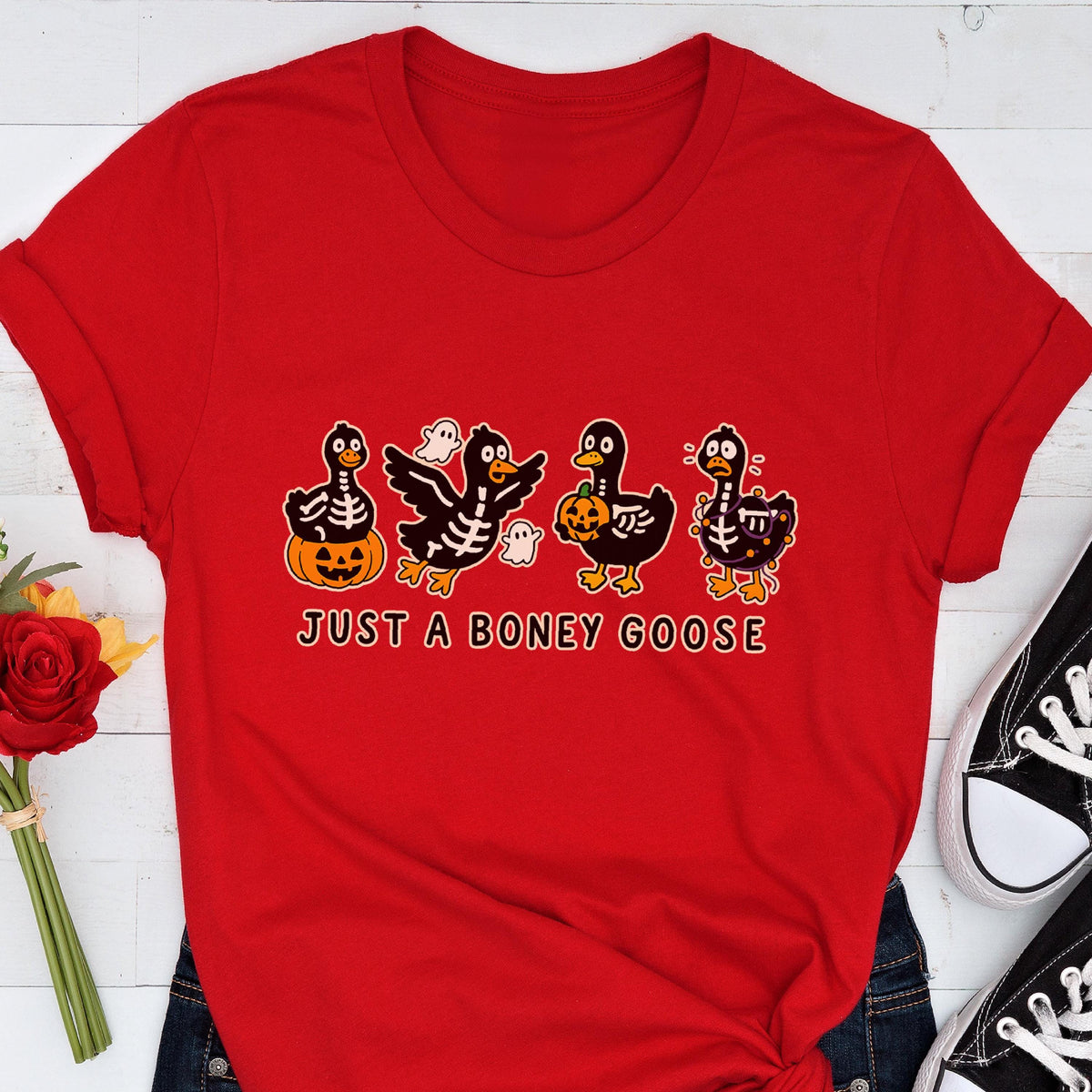 May include: Red t-shirt featuring a Halloween-themed graphic. The design includes four cartoon geese dressed as skeletons, a jack-o'-lantern, and ghosts. The text "JUST A BONEY GOOSE" is printed below the geese.