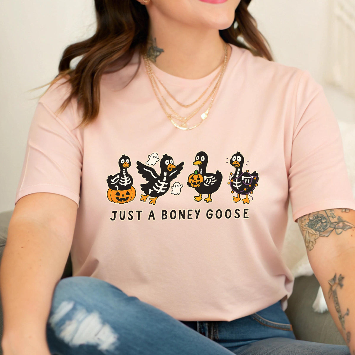 May include: A light pink t-shirt featuring a Halloween-themed graphic. The design includes four black geese with skeleton details, holding pumpkins and ghosts. The text "JUST A BONEY GOOSE" is printed below the geese.