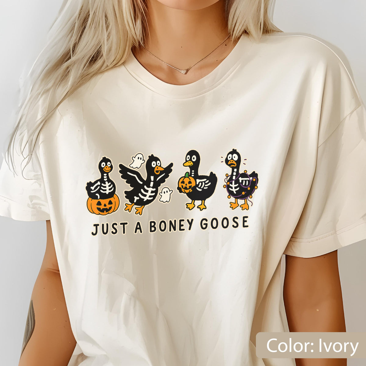 May include: Ivory-colored t-shirt featuring a Halloween-themed graphic. The design includes four cartoon geese with skeleton details, pumpkins, and ghosts. The text "JUST A BONEY GOOSE" is printed below the geese.