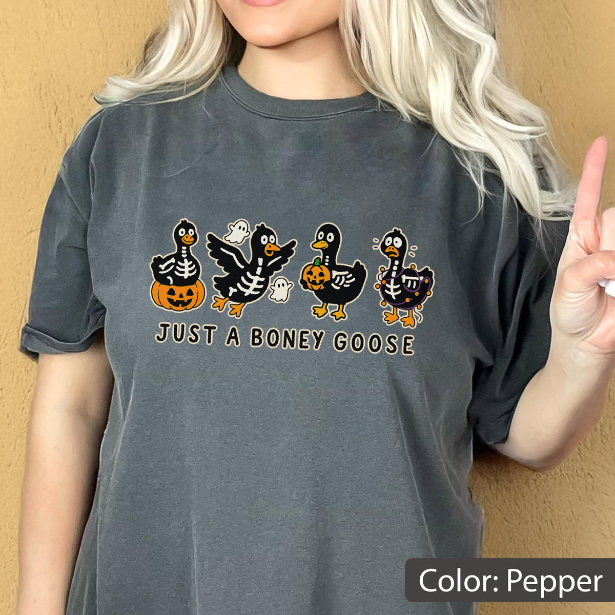May include: Pepper-colored t-shirt featuring a Halloween-themed graphic. The design includes four cartoon geese with skeleton details, pumpkins, and ghosts. The text "JUST A BONEY GOOSE" is printed below the geese.