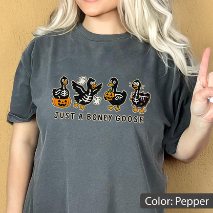 May include: Pepper-colored t-shirt featuring a Halloween-themed graphic. The design includes four cartoon geese with skeleton details, pumpkins, and ghosts. The text "JUST A BONEY GOOSE" is printed below the geese.