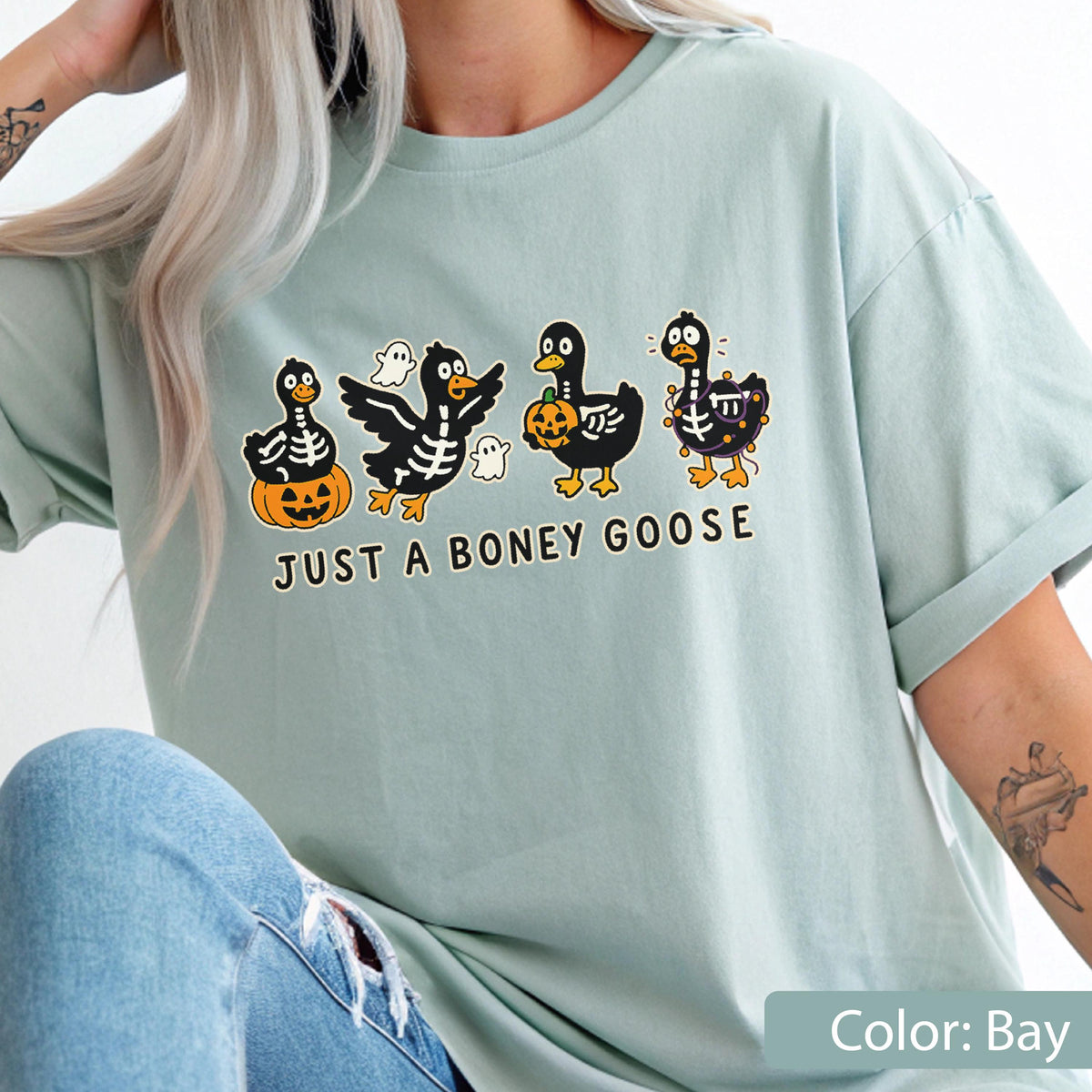 May include: A light blue t-shirt featuring a Halloween-themed graphic. The design includes four cartoon geese with skeleton details, holding pumpkins and accompanied by ghosts. The text "JUST A BONEY GOOSE" is printed below the geese.